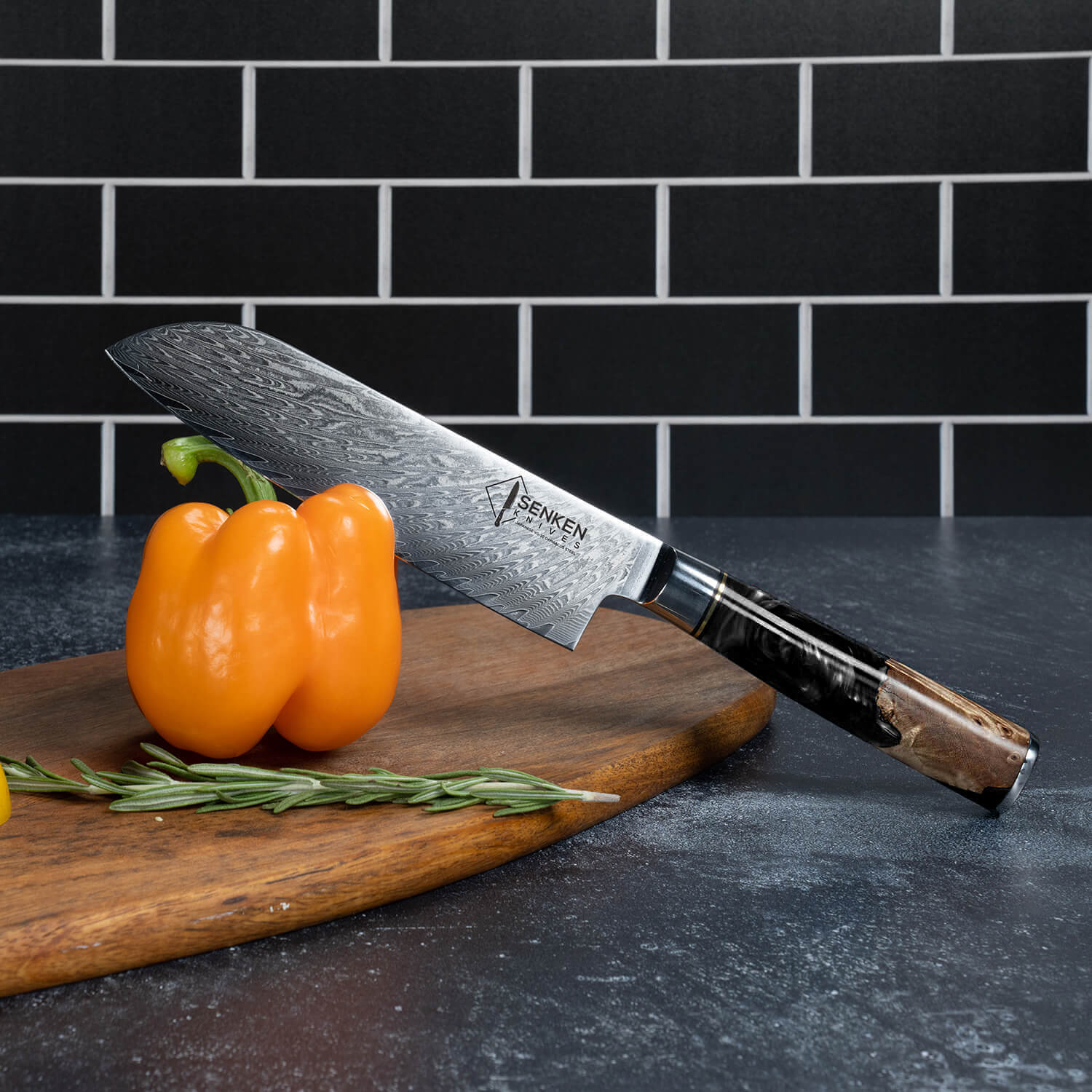 Knife with a black wooden handle on a cutting board next to two orange peppers and a sprig of rosemary against a black tiled wall.