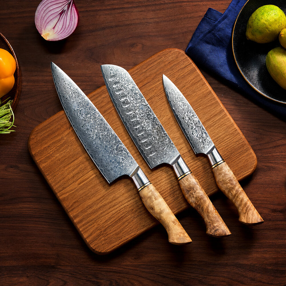 Three kitchen knives with patterned steel blades and light wood handles arranged on a wooden cutting board over a dark wood table, with food props around them.