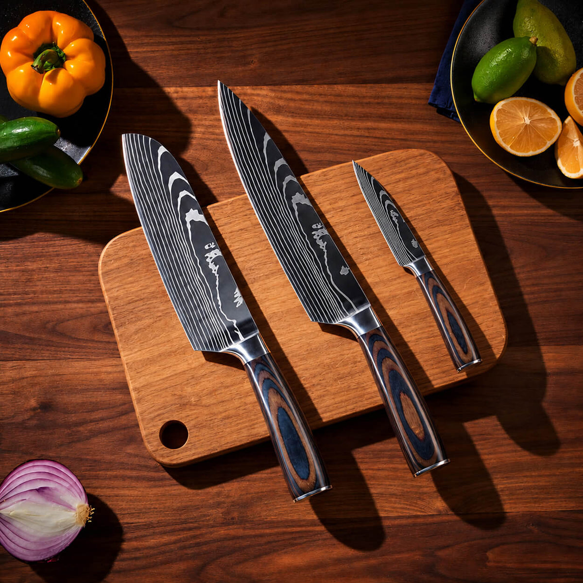 Three kitchen knives with patterned steel blades and dark wood handles arranged on a wooden cutting board over a wooden table, with vegetables around the edges.
