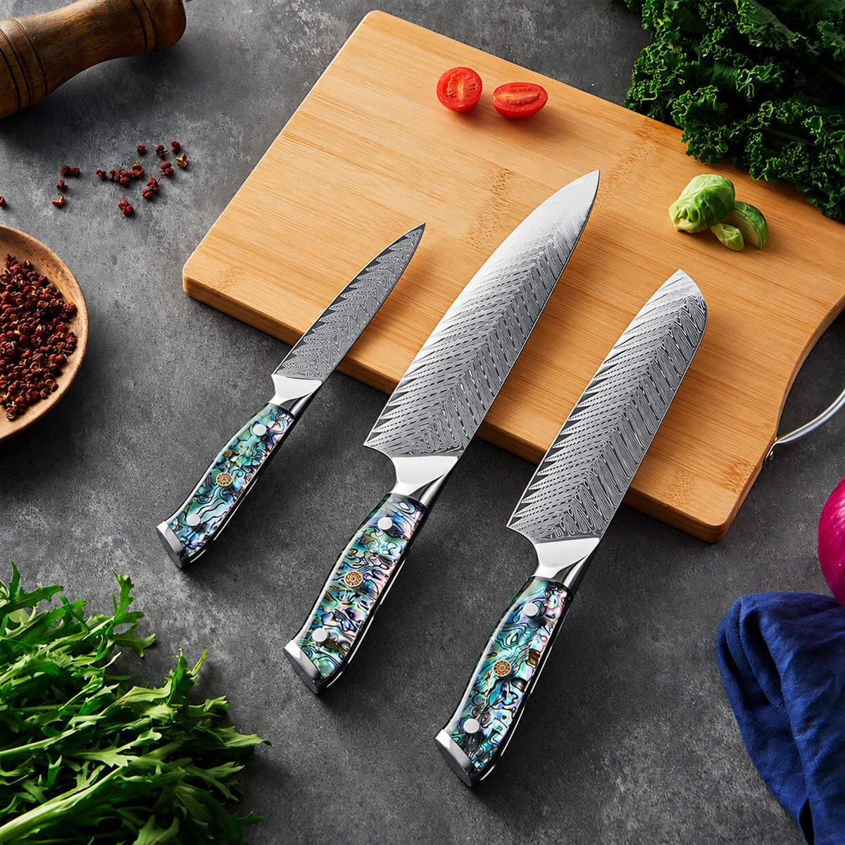 Three kitchen knives with patterned steel blades and colorful mosaic handles are arranged on a gray surface beside a wooden cutting board with tomatoes, Brussels sprouts, and herbs.