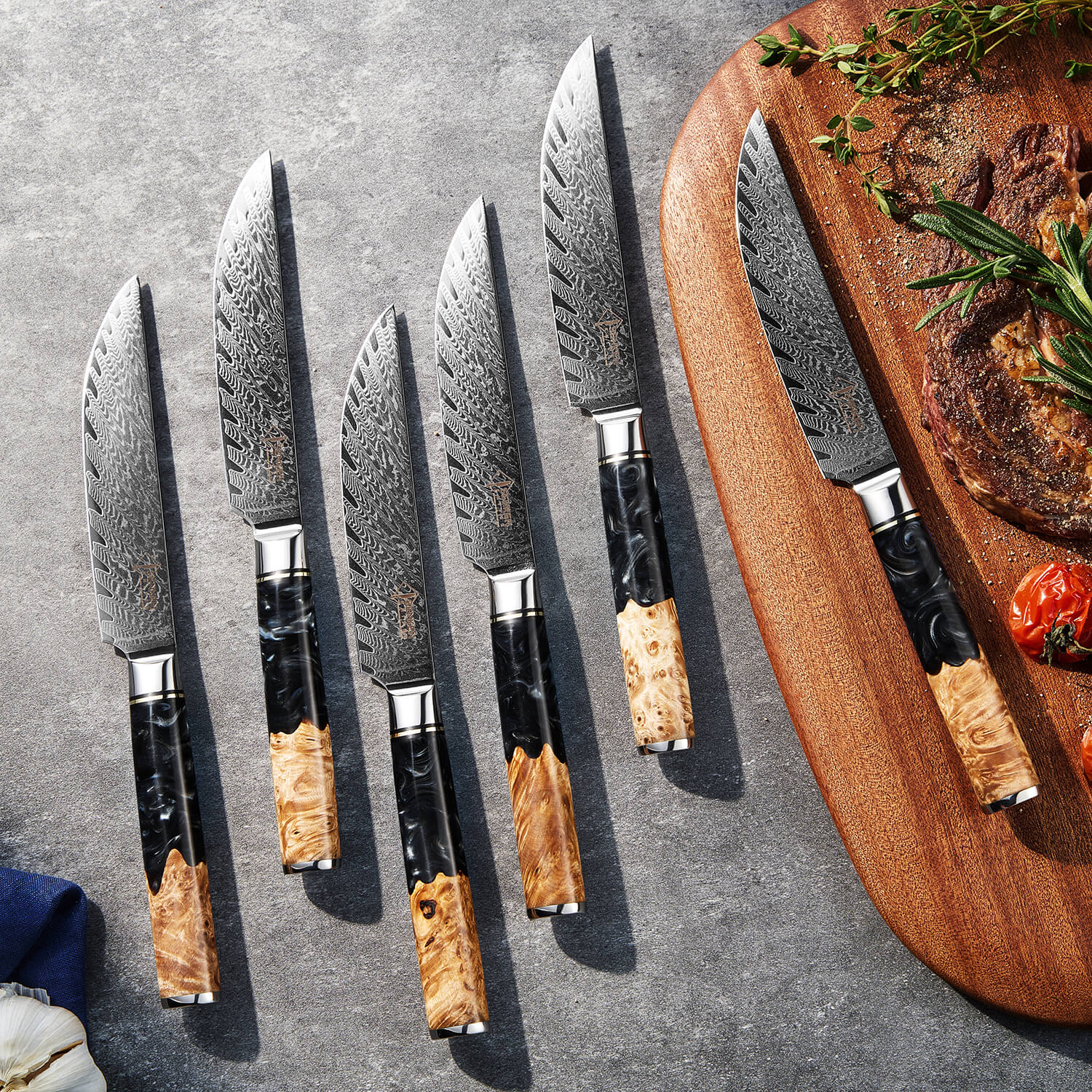 Set of knives with wooden handles on a gray surface, next to a wooden cutting board with food.