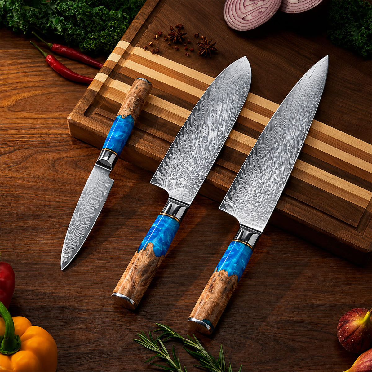 Three kitchen knives with patterned steel blades and blue-and-brown wood handles arranged on a wooden cutting board and dark wood table, surrounded by food props like peppers, parsley, onions, and figs.